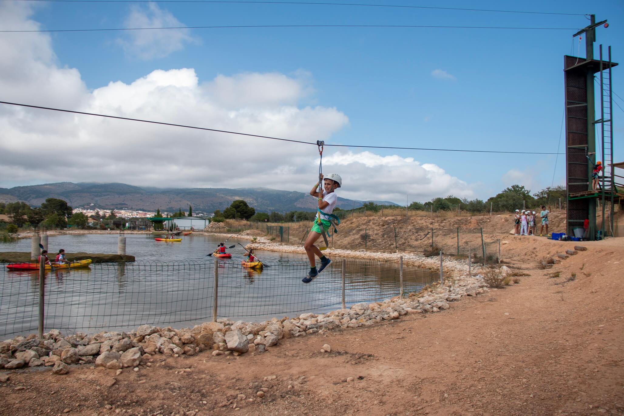 joven en tirolina sobre lago con kayaks al fondo